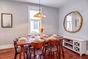 A dining area with a wooden table set for six, a hanging pendant light, a white console with board games, and a large mirror on the wall. - Hot Tub, Newly Updated, Near DT, Free parking | TS (Peterborough)