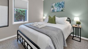 A bedroom with a bed, two bedside tables with lamps, a window with blinds, and a wall-mounted painting. - Cozy & Comfortable, Downtown, Parking | TJSI (Burlington)