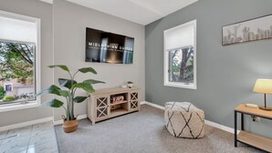 A modern living room with a grey wall, a mounted TV, a wooden console table, a potted plant, a cushioned ottoman, and a side table with a lamp. - Cozy & Comfortable, Downtown, Parking | TJSI (Burlington)