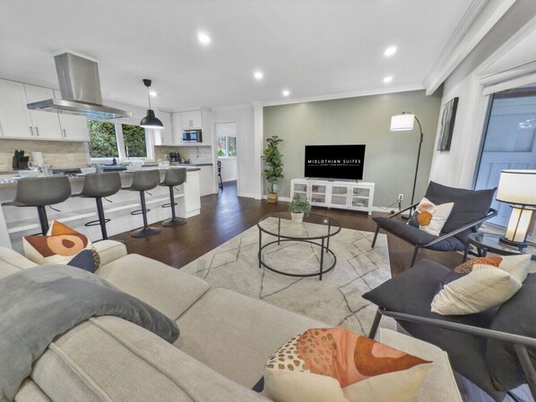 A modern living room with a sofa, armchairs, and a coffee table. There is a kitchen area with white cabinets and a stainless steel range hood. A flat-screen TV is mounted on the wall, and a potted plant is on a side table.