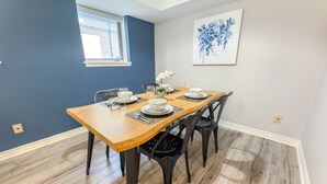 A dining area with a wooden table set for four, black chairs, a window, and a blue floral painting on the wall. - Downtown, 2 Bedroom, Parking | TJS3 (Burlington)