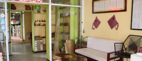 A room with a wooden coffee table, a white sofa, a black chair, and a fan. There is a glass door leading to a shop with a neon sign.