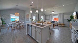 A spacious kitchen with a central island, dining area with chairs, and a living room with a sofa and chairs. - Two Port A Homes Near Beach & Nature Preserve Ideal Group Retreat (Port Aransas)