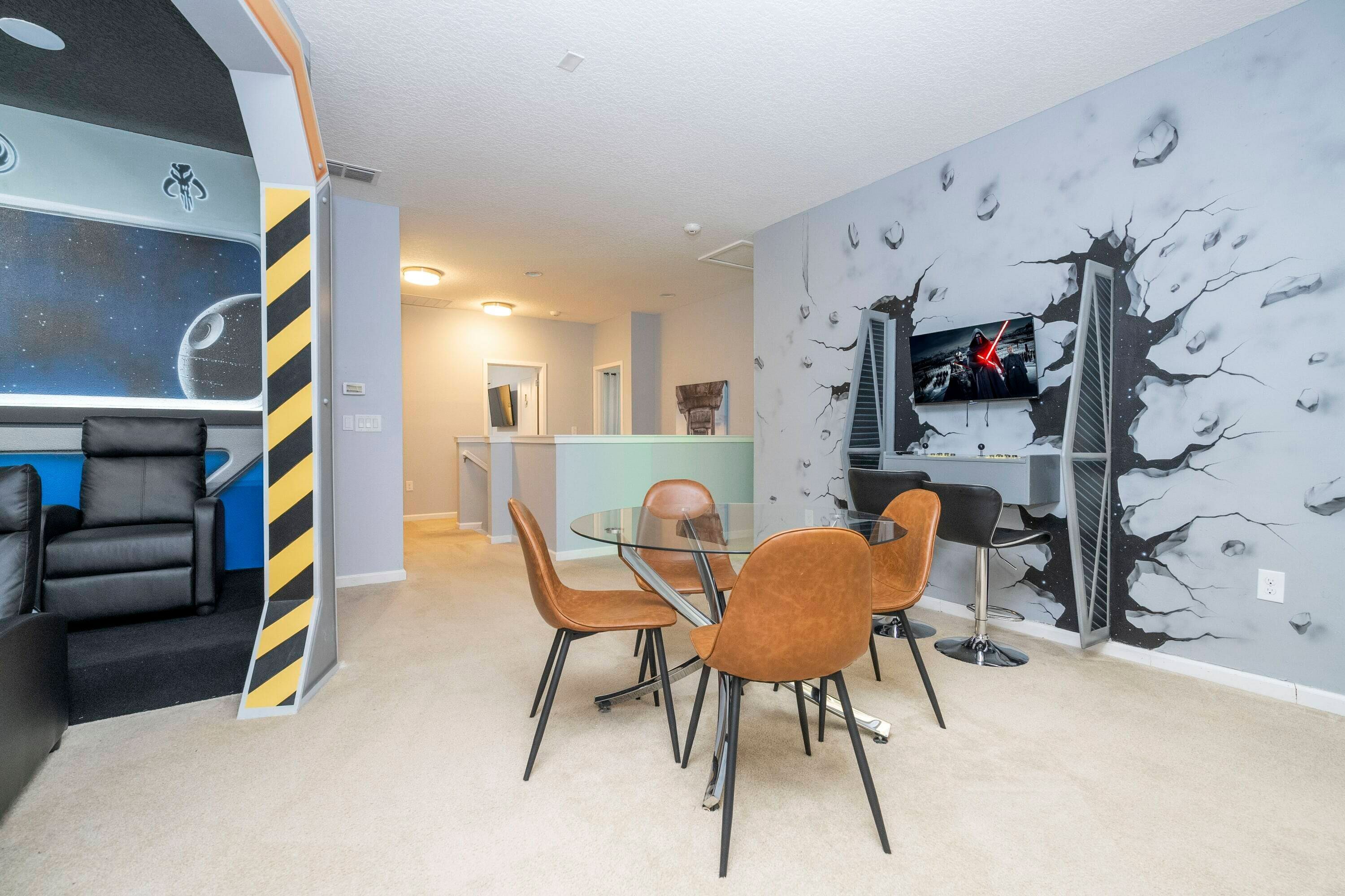 Un salon moderne avec une table et des chaises en verre, un canapé, un téléviseur et un mur orné d’une fresque sur le thème de l’espace.