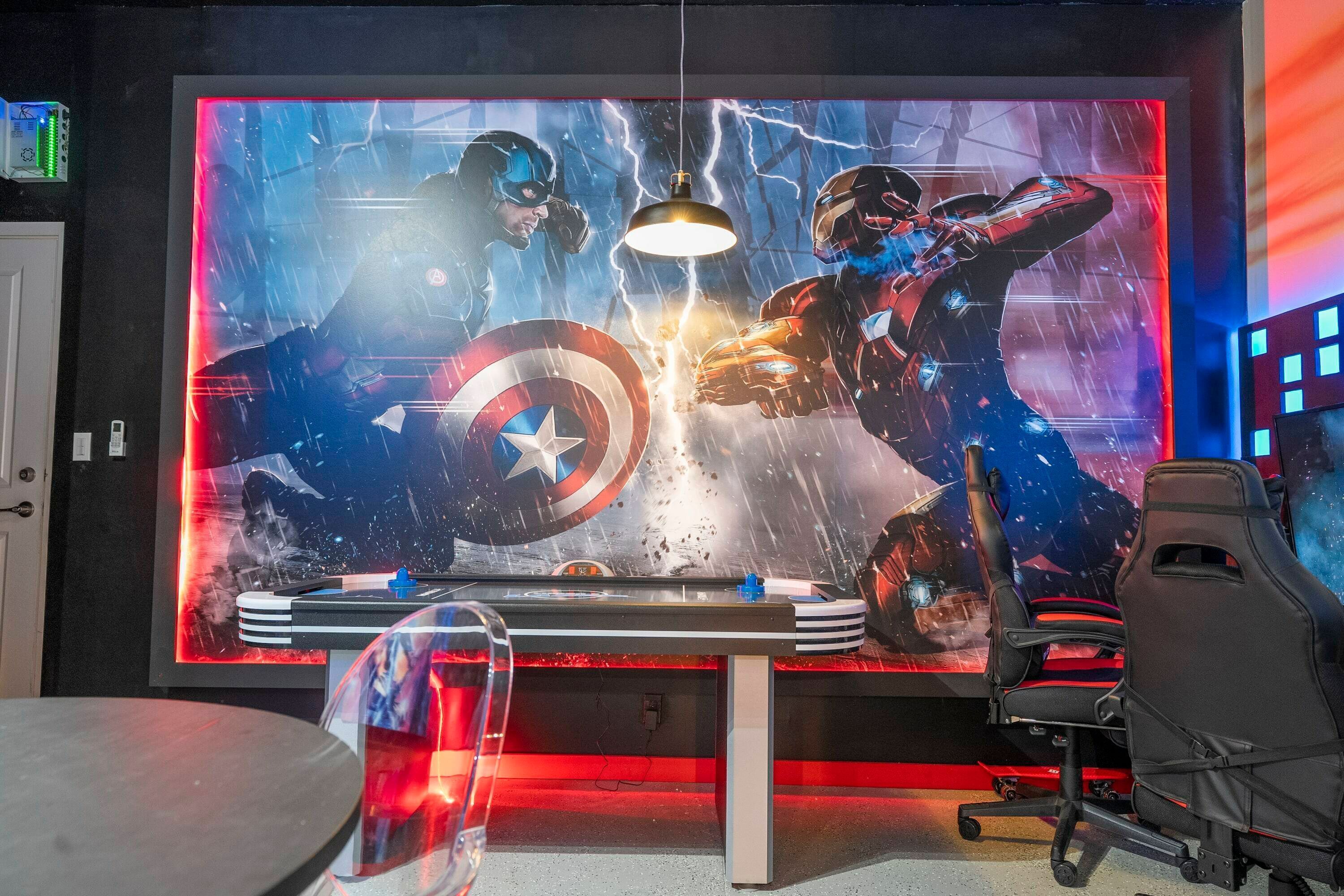 A gaming setup with a large screen displaying a superhero battle scene, a desk, and gaming chairs.