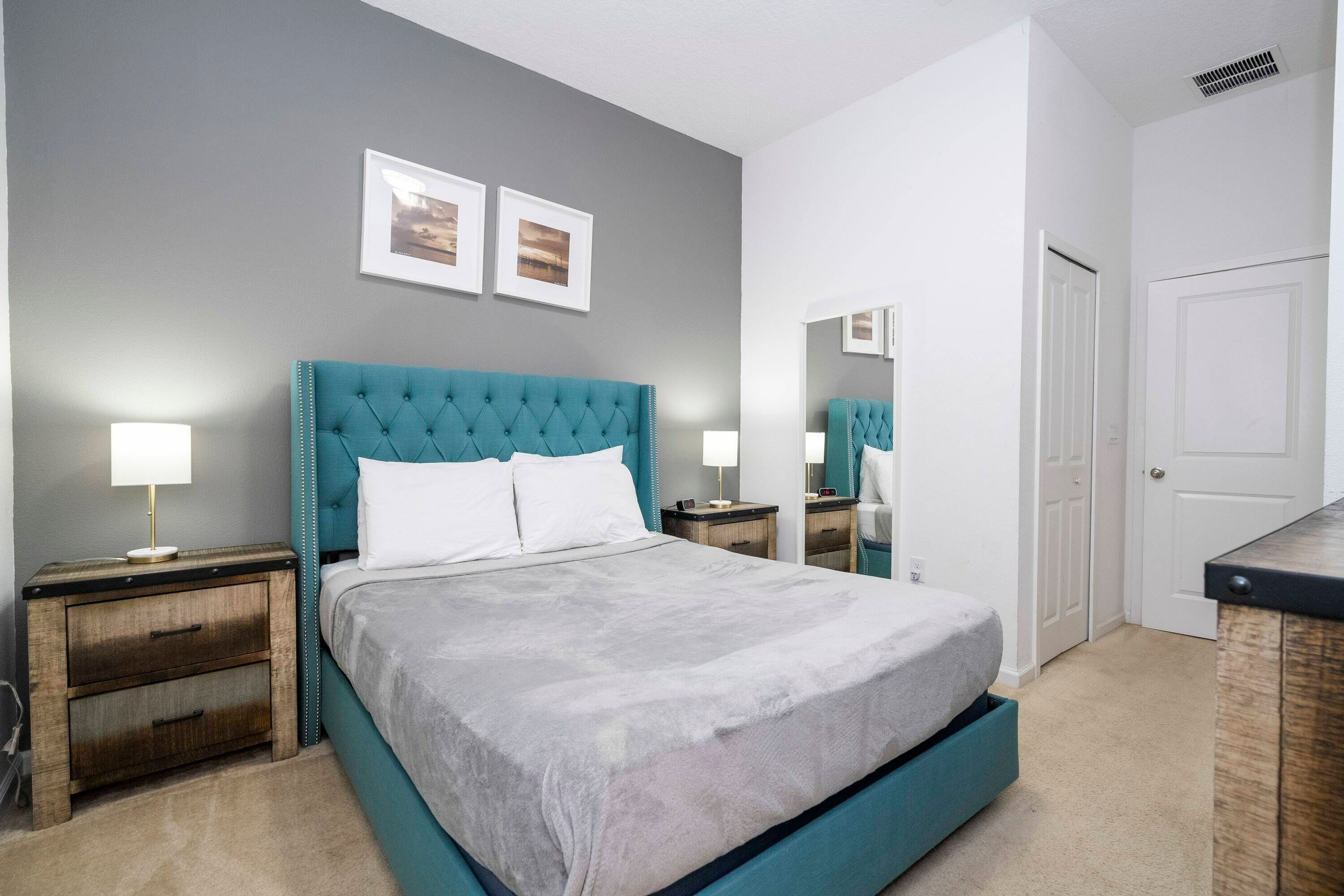 A bedroom with a teal upholstered headboard, a large bed with a grey bedspread, two bedside tables with lamps, and a door with a mirror.