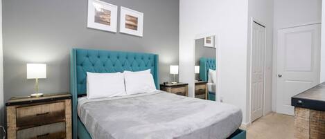 A bedroom with a teal upholstered headboard, a large bed with a grey bedspread, two bedside tables with lamps, and a door with a mirror.