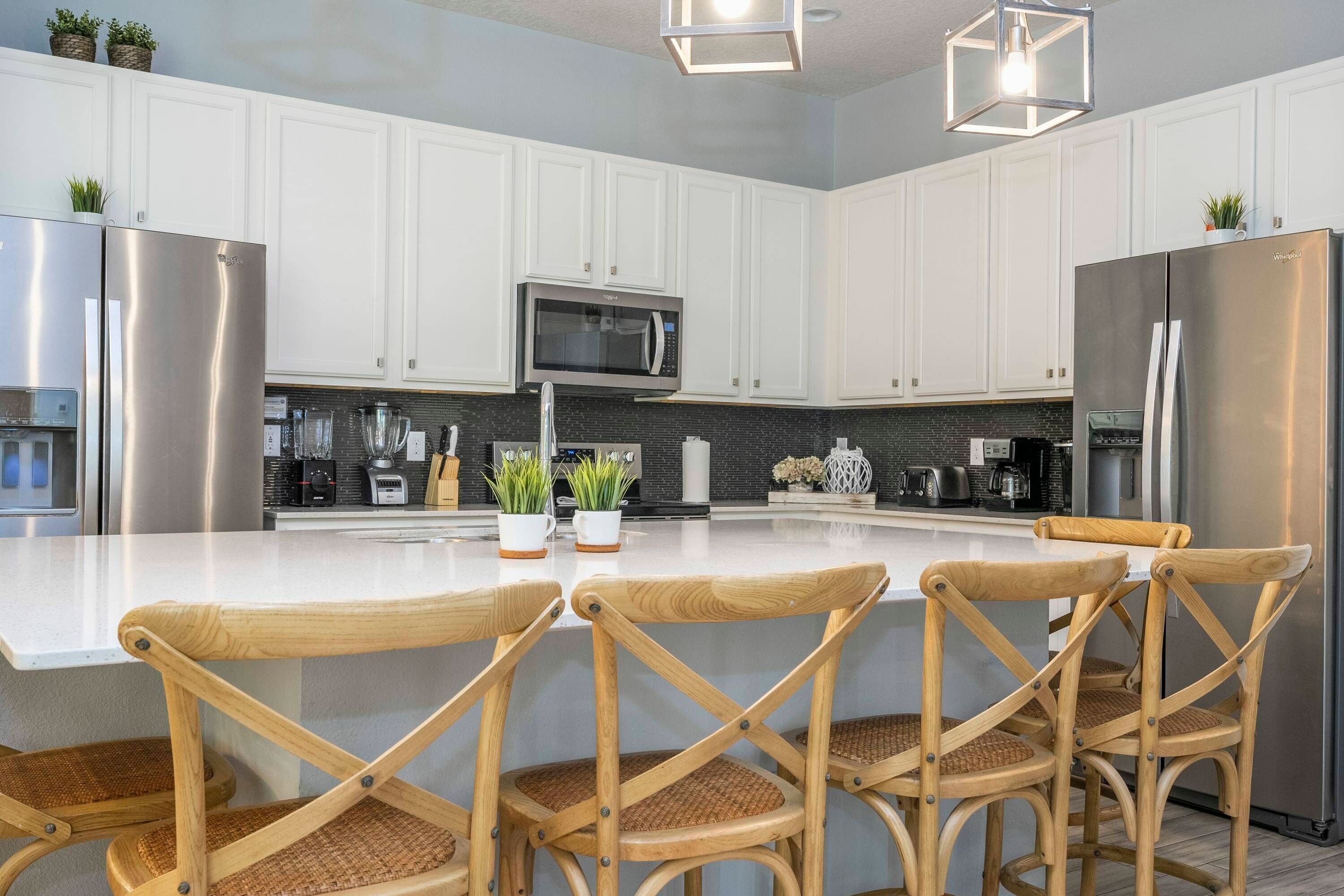 A modern kitchen with white cabinets, stainless steel appliances, a central island with chairs, and decorative plants.