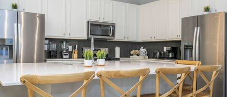 A modern kitchen with white cabinets, stainless steel appliances, a central island with chairs, and decorative plants.