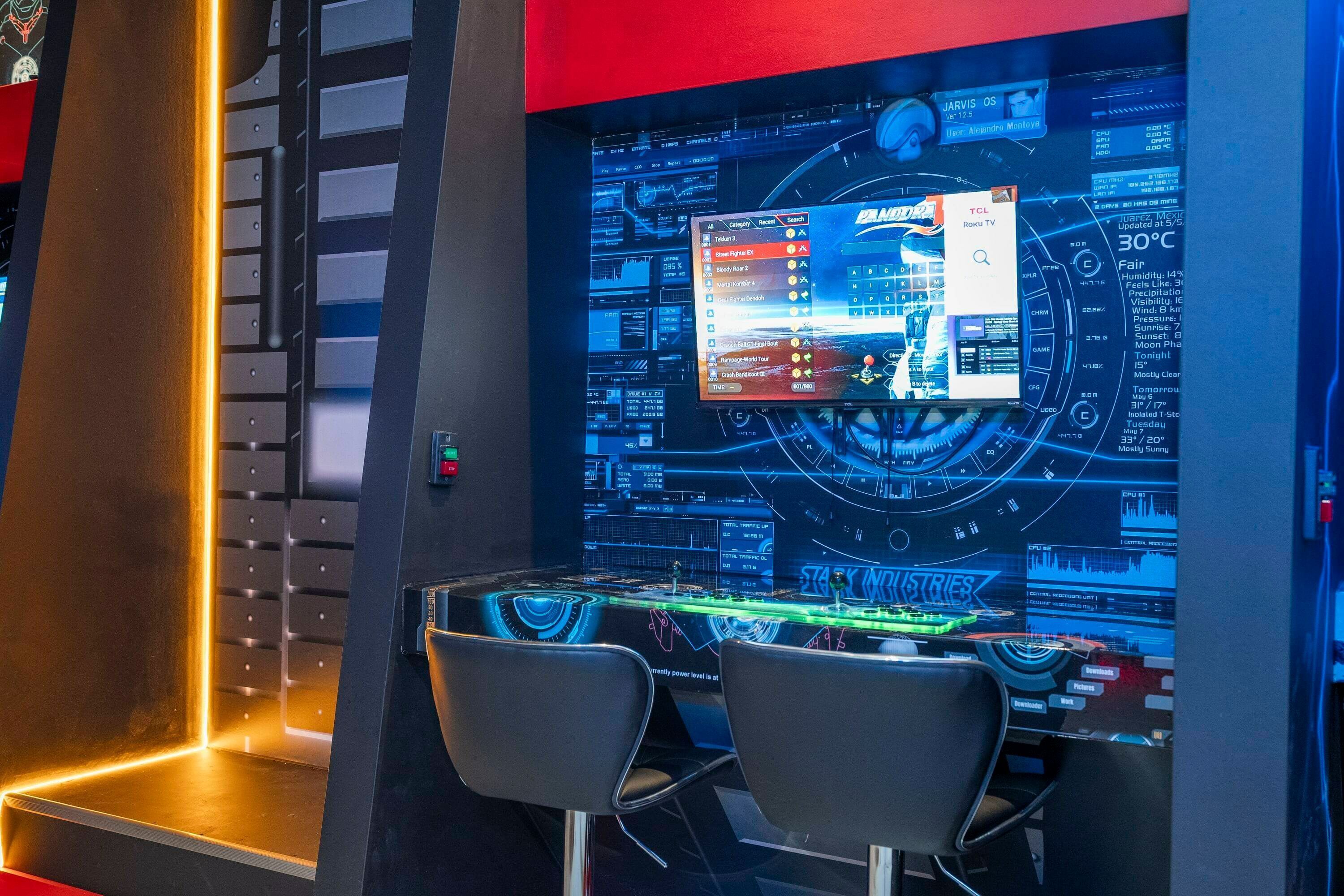 A gaming setup with a large screen displaying a game interface, two chairs, and a control panel.