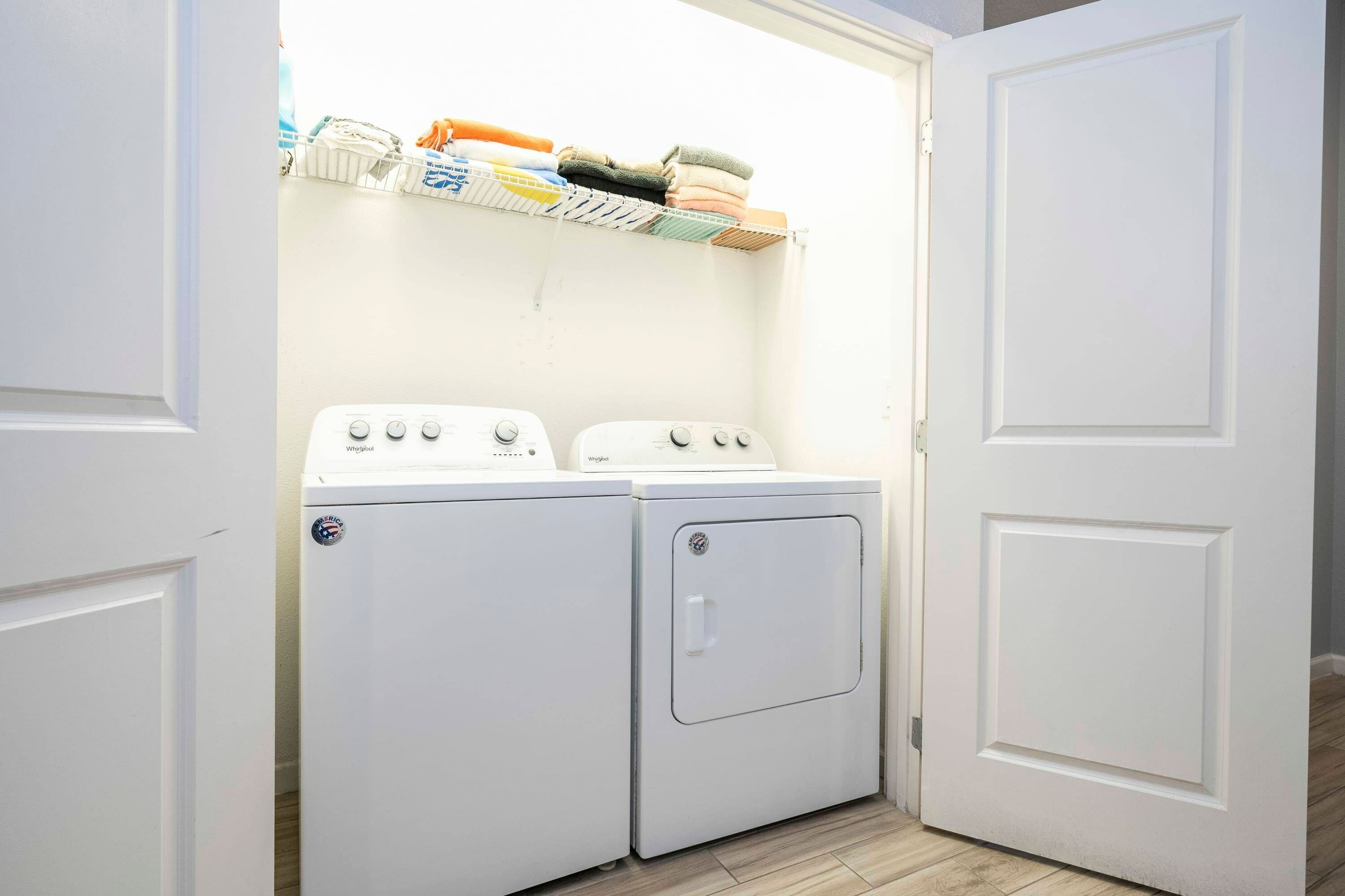 A laundry room with two white washing machines and a shelf above them holding folded clothes.
