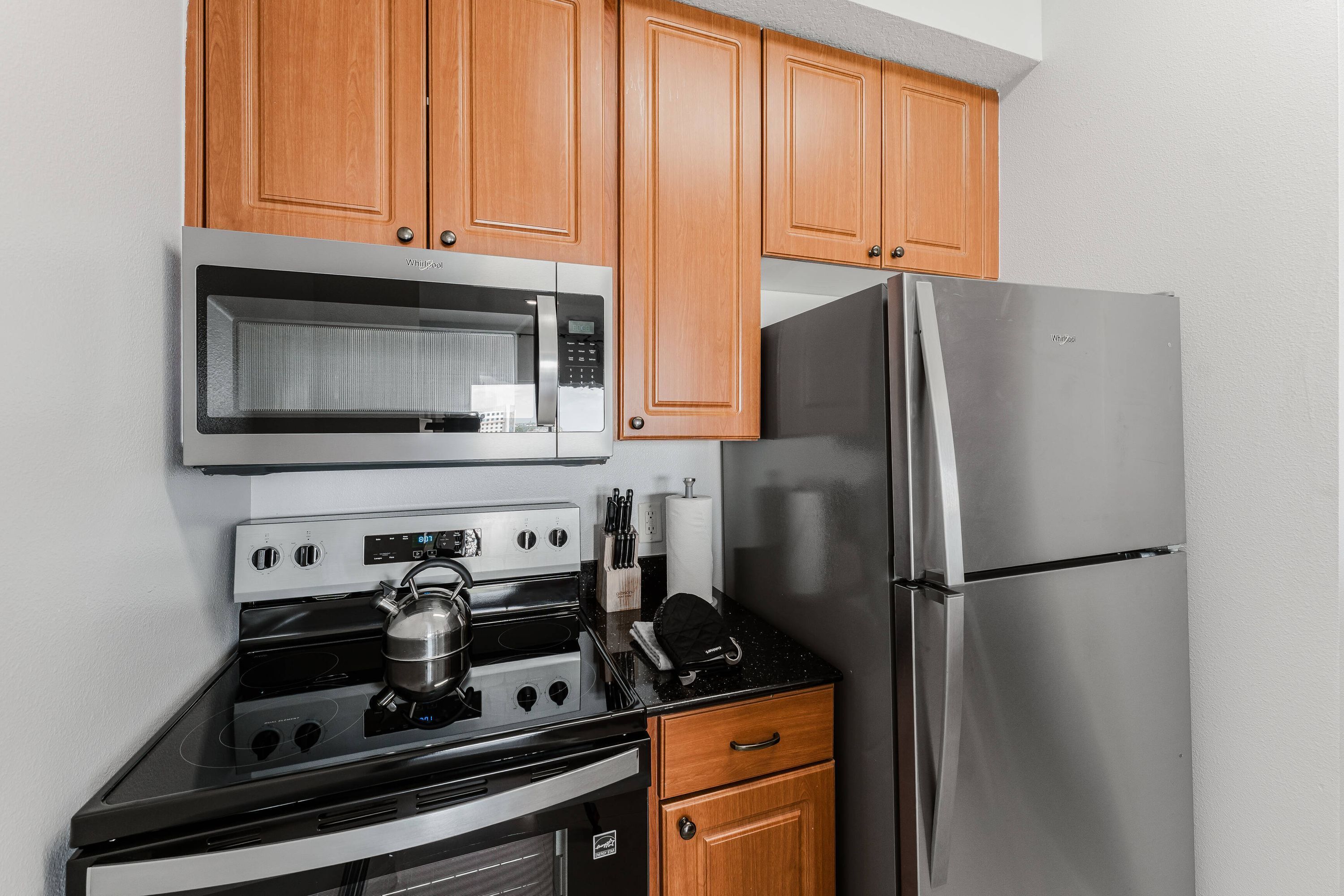 A kitchen with wooden cabinets, a stainless steel refrigerator, a microwave, a stove, and a kettle.