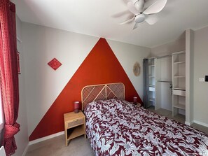 Ein Schlafzimmer mit geometrischem Wanddesign, einem Bett mit floralem Überwurf, einem Nachttisch mit Lampe und einem Kleiderschrank.