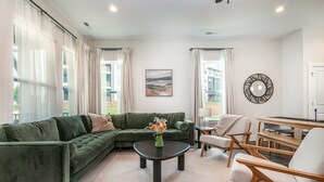 A modern living room with a green sectional sofa, a round coffee table with a vase of flowers, a dining table with chairs, and large windows with curtains. - UNCC 4BR | Pet-Friendly Stay (Charlotte)