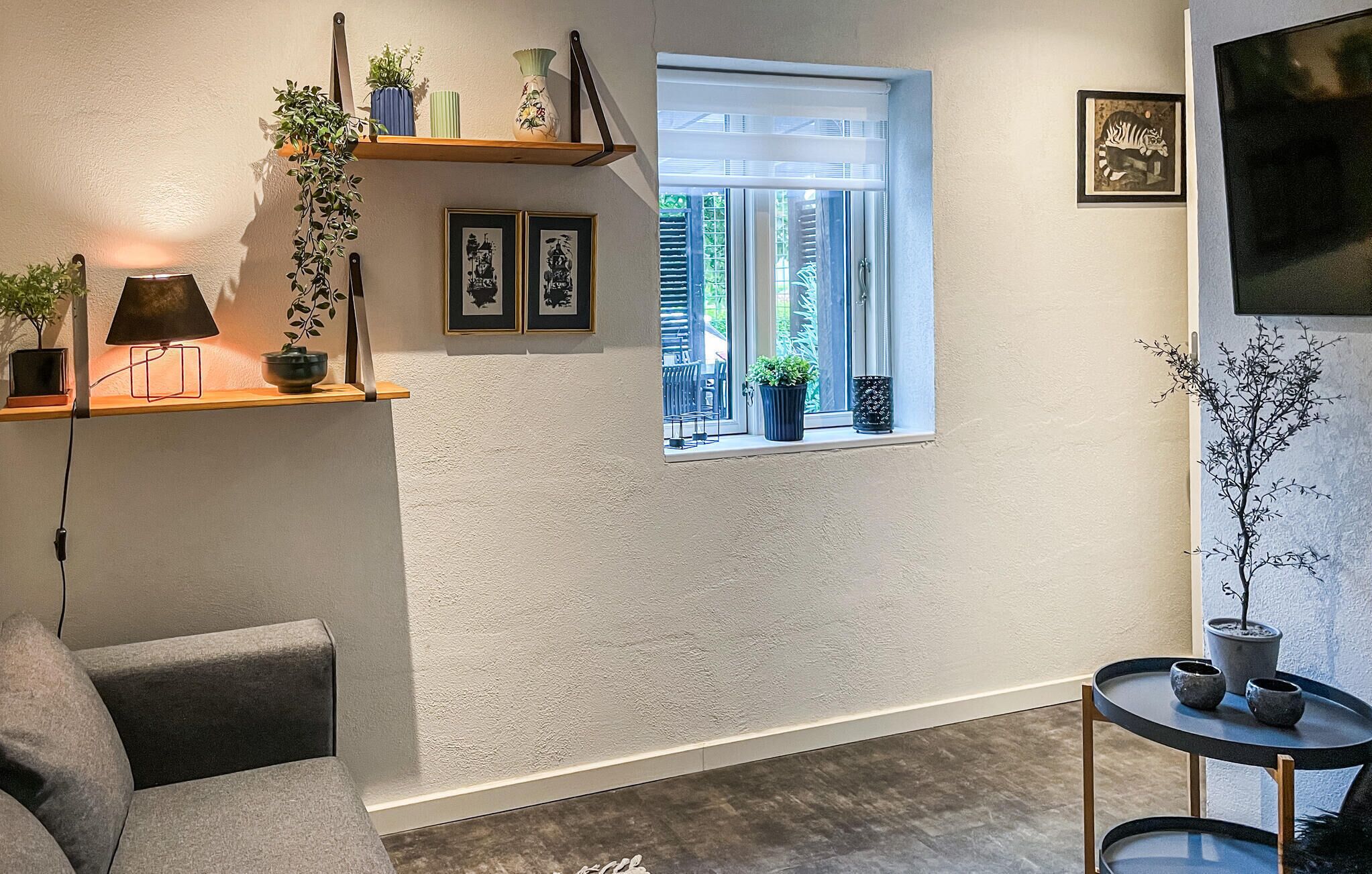 Un salon moderne avec un canapé gris, une petite table ronde, une fenêtre avec des stores et divers objets décoratifs disposés sur des étagères.