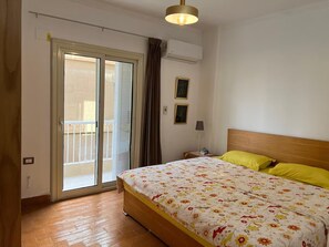 Un dormitorio con una cama, una mesita de noche, una lámpara, un equipo de aire acondicionado y un ventanal corredero que da a un balcón.