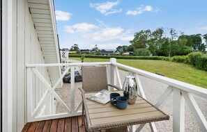 A wooden deck with a wicker chair, a table with two mugs and a book, and a view of a green landscape with houses in the distance. - Stunning home in Brenderup Fyn (Brenderup Fyn)