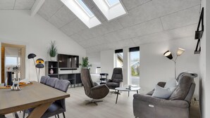 A modern living room with a dining area, grey sofas, a TV, and a kitchen in the background.