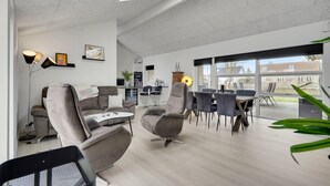 A spacious living area with modern furniture, a kitchen in the background, and large windows offering a view of the outside.