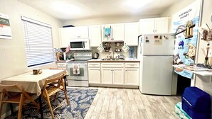 A compact kitchen with white cabinets, a microwave, a stove, and a refrigerator. There is a dining table with chairs and a patterned rug.