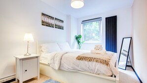 A bedroom with a bed, bedside table, lamp, window, and artwork on the wall.