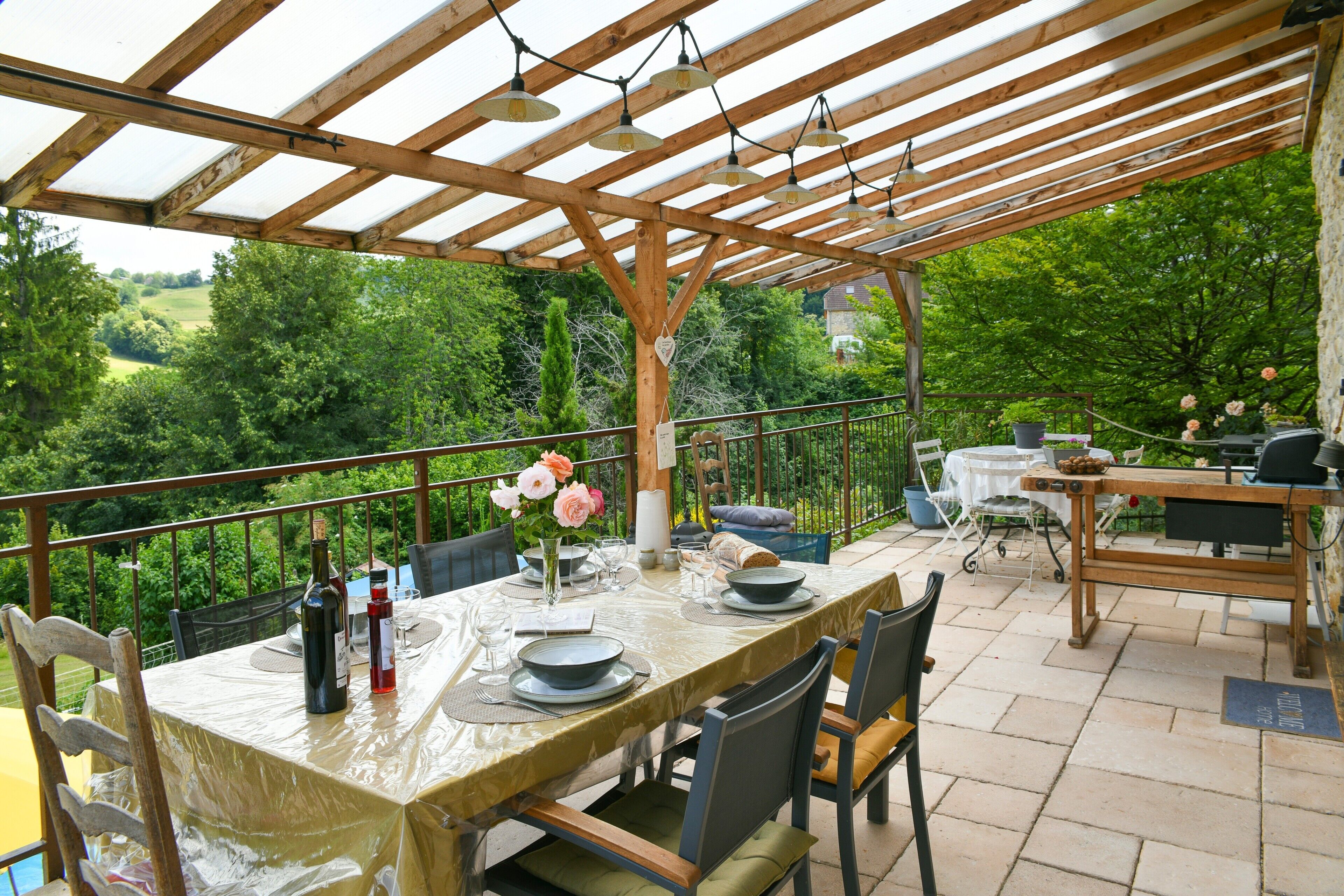 Een terras buiten met een overdekte houten eettafel, omringd door stoelen en uitzicht op groen.