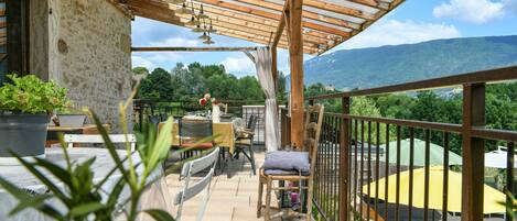 A covered outdoor patio with a table and chairs, a potted plant, and a view of a mountainous landscape.