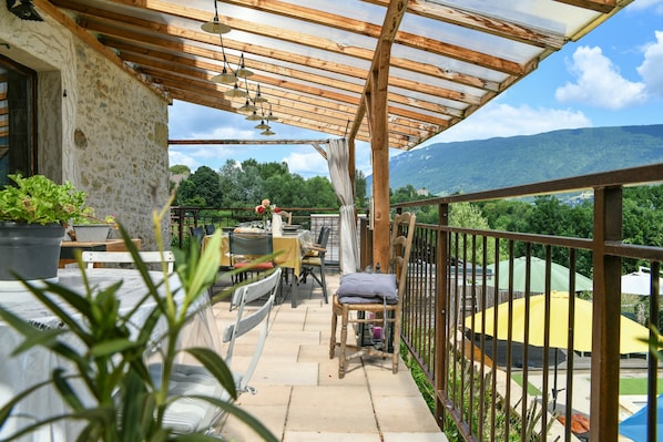 A covered outdoor patio with a table and chairs, a potted plant, and a view of a mountainous landscape.