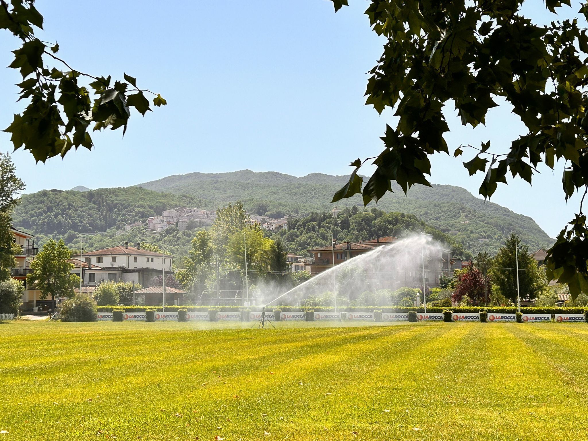 A grassy field with a fountain in the center, surrounded by buildings and a hill in the background.