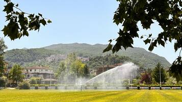 A grassy field with a fountain in the center, surrounded by buildings and a hill in the background.