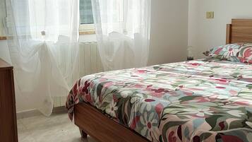 Une chambre à coucher avec un lit, une tête de lit en bois, une table de chevet et une fenêtre ornée de rideaux légers.