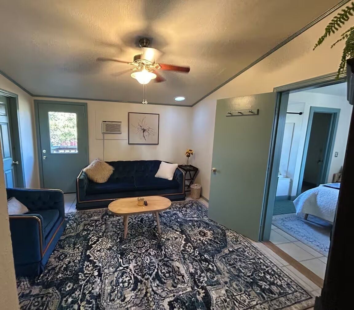 A living room with a dark blue sofa, a patterned rug, a ceiling fan, and a view into a bedroom with a bed.