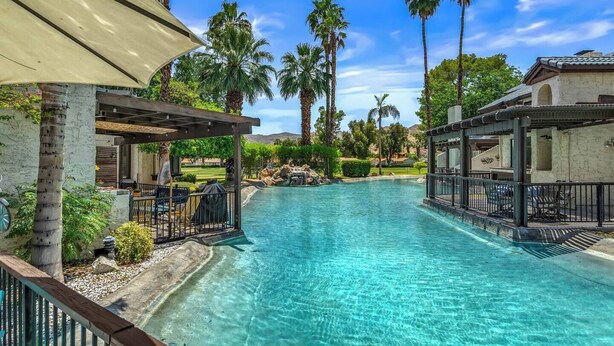 A clear pool with a pergola and seating area, surrounded by palm trees and a stone building.