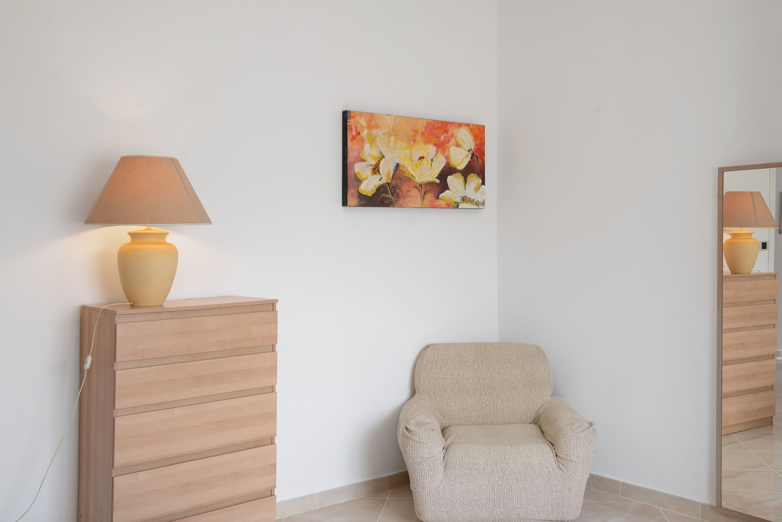Una stanza con una poltrona beige, un armadietto in legno, una lampada e un quadro appeso al muro.