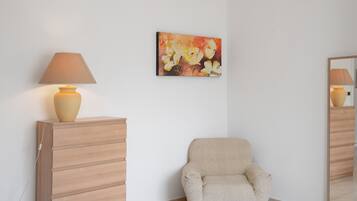 Una stanza con una poltrona beige, un armadietto in legno, una lampada e un quadro appeso al muro.