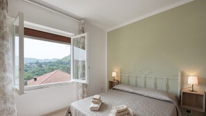 A bedroom with a bed, a window with curtains, a nightstand with a lamp, and a view of a green landscape. - Villa 'Chiara' with Mountain View and Private Terrace (Lumarzo)