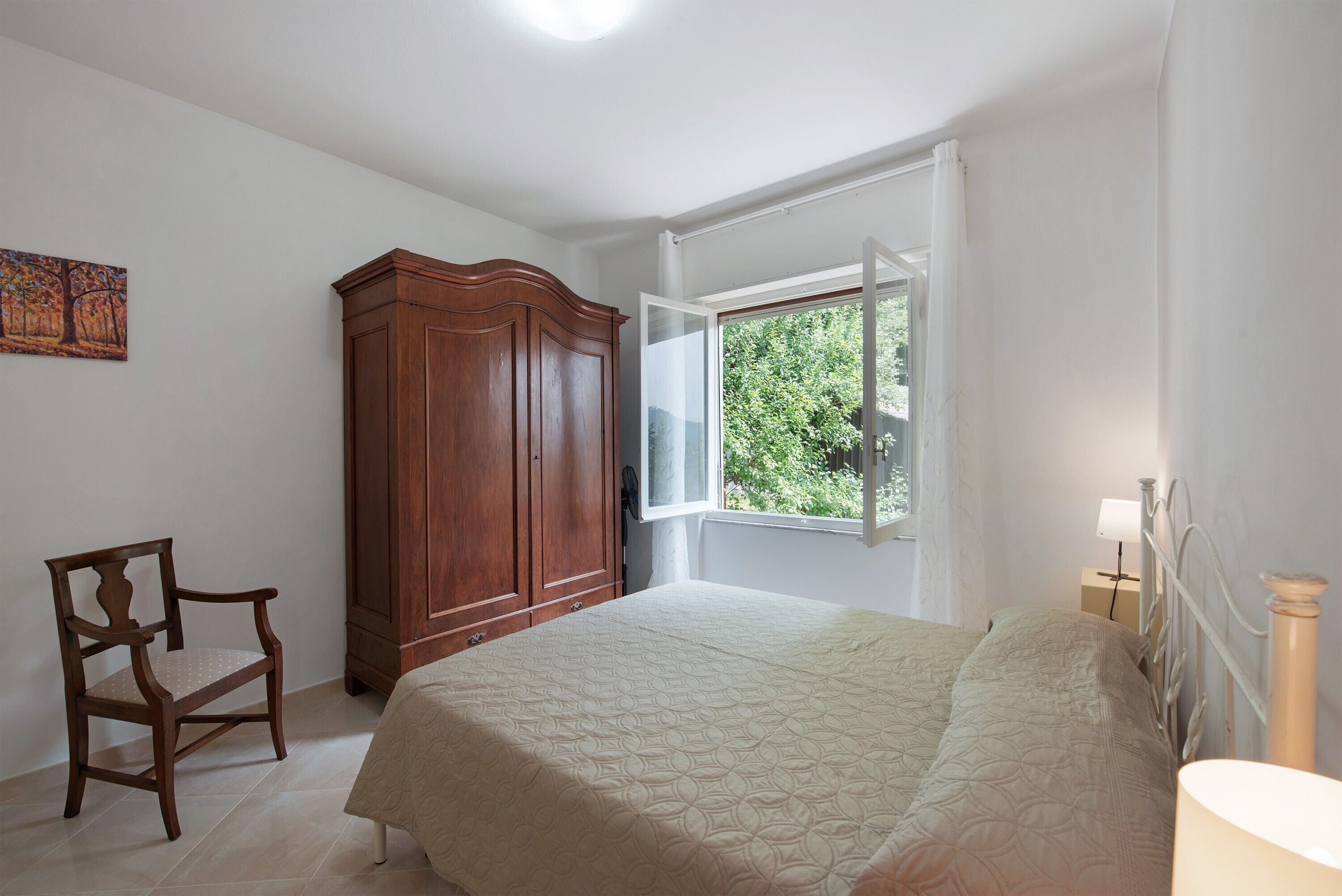 Una camera da letto con un letto, un armadio in legno, una sedia, una finestra con tende e una lampada a parete.