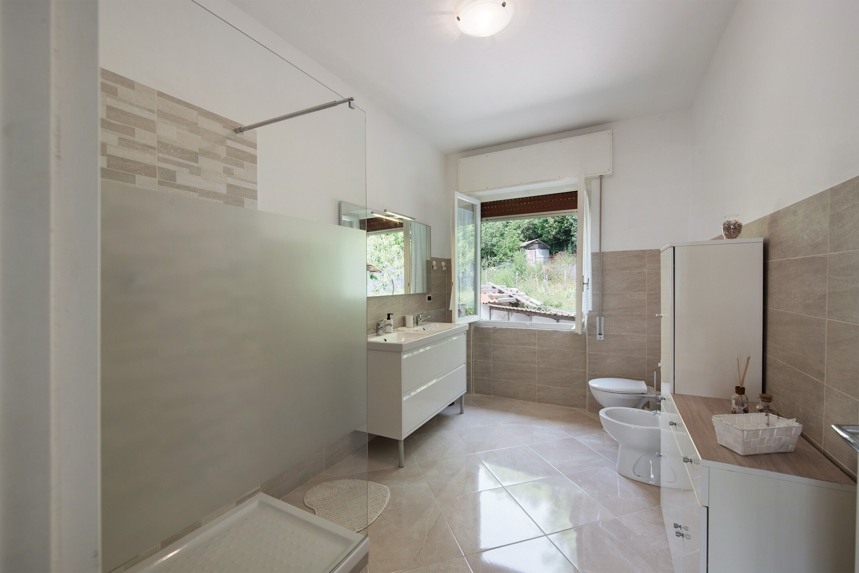 Een moderne badkamer met douche, toilet en wastafel, met een grote spiegel en uitzicht op het buitengebied.