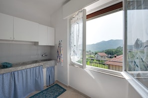 A kitchen with white cabinets, a window with a view of houses and mountains, and a blue patterned rug.
