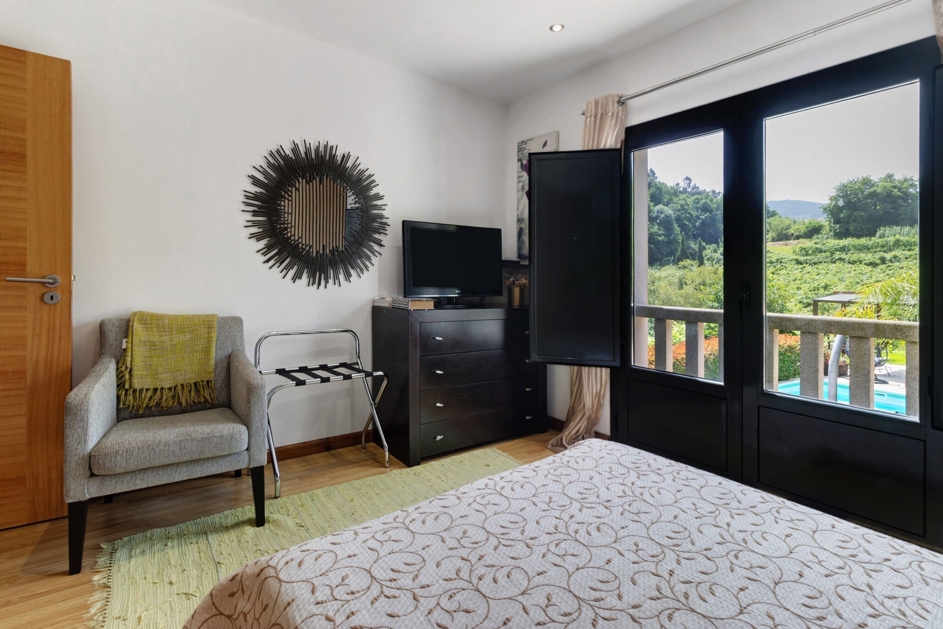 Een slaapkamer met een bed, een stoel, een televisie, een dressoir en een balkon met uitzicht op groen.