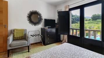 Een slaapkamer met een bed, een stoel, een televisie, een dressoir en een balkon met uitzicht op groen.