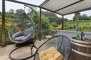 A covered outdoor seating area with a hanging chair, a cushioned chair, and a wooden barrel with bottles. - Holiday Home 'Cevidade' with Lake View, Wi-Fi and Air Conditioning (Melgaço)