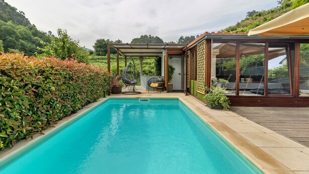 Une piscine avec une terrasse en bois, un espace salon couvert et une vue sur une colline verdoyante.