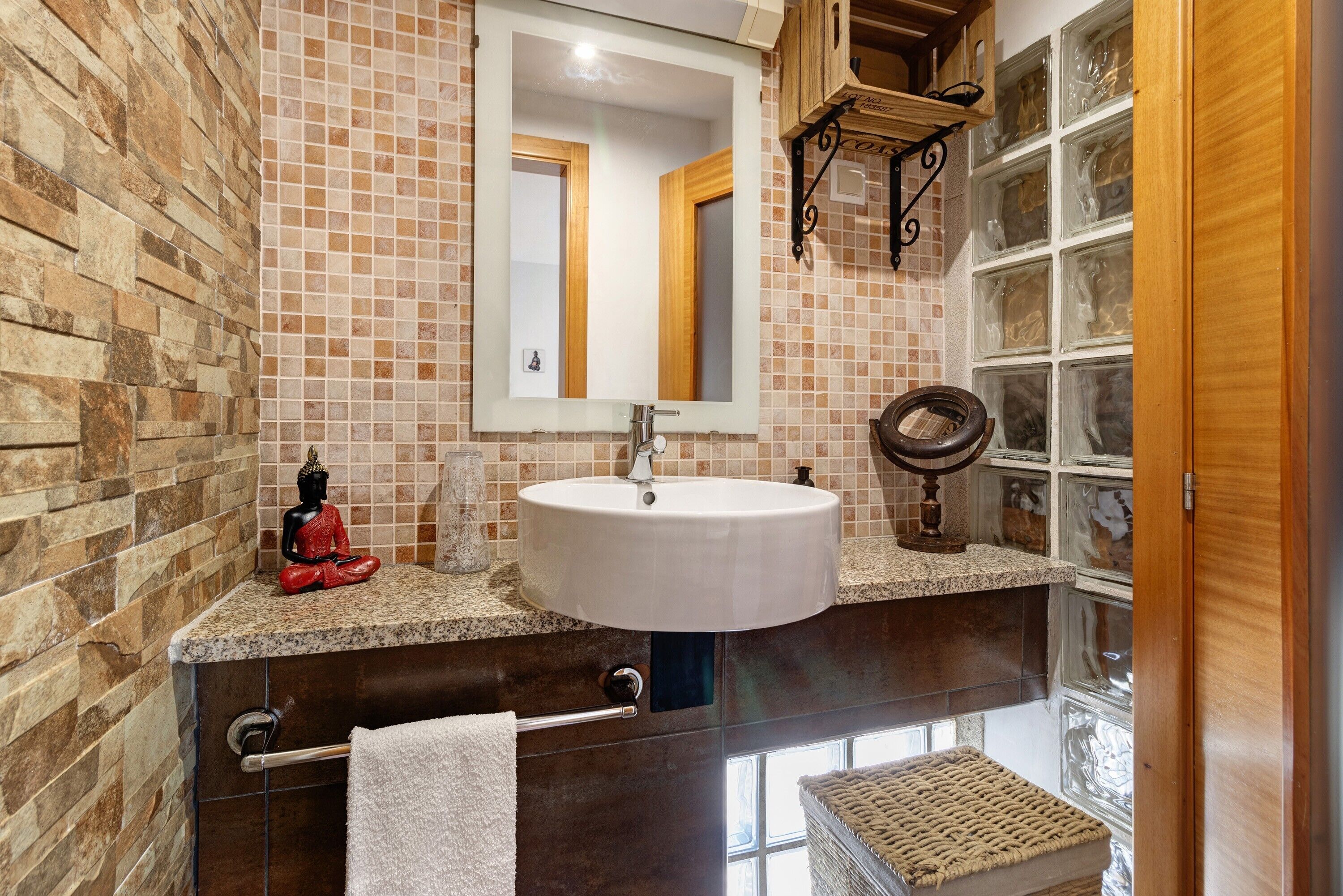 A bathroom with a stone wall, a white sink, a mirror, a Buddha statue, and a window with a view.