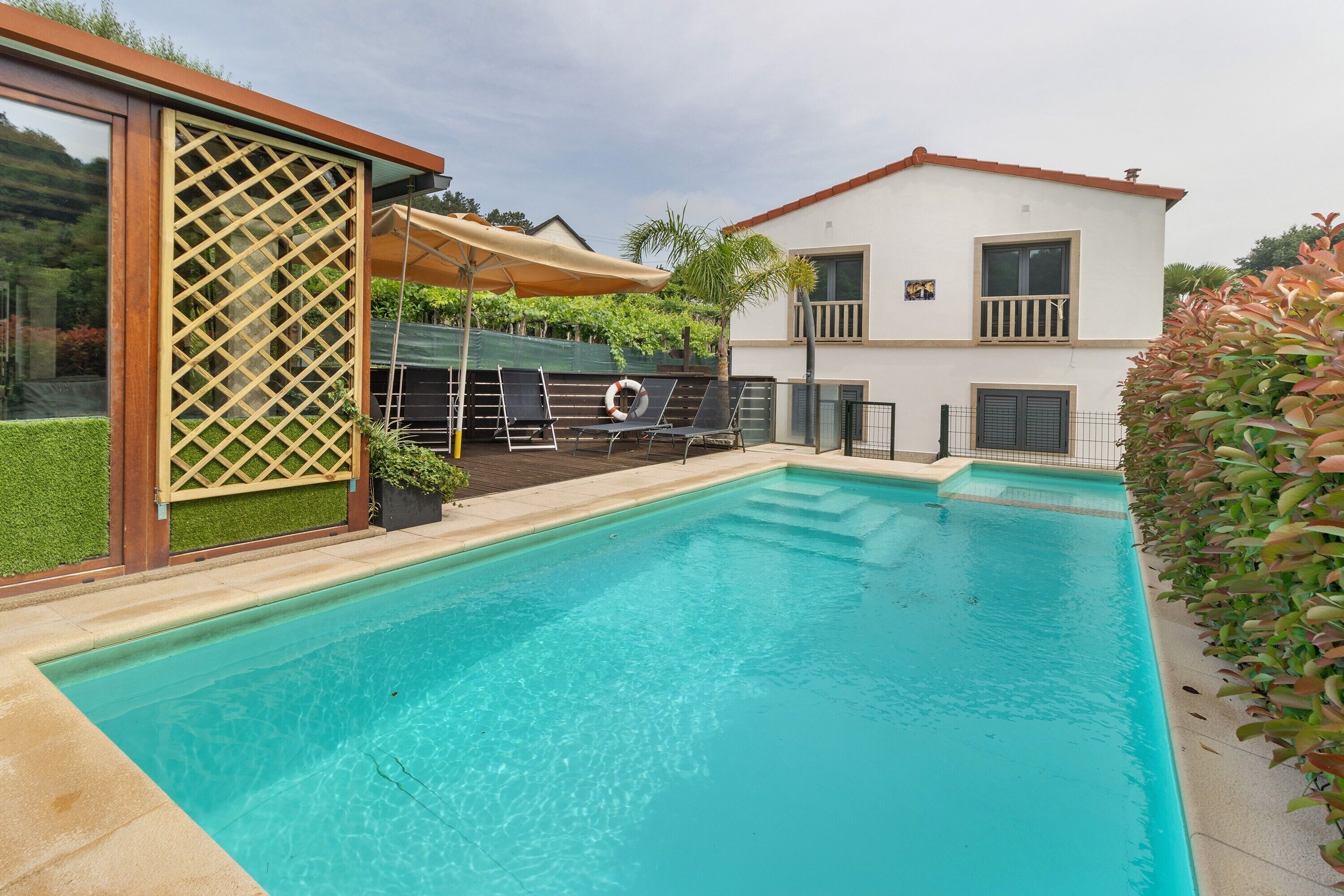 A swimming pool with a wooden deck, a covered seating area, and a two-story building in the background.