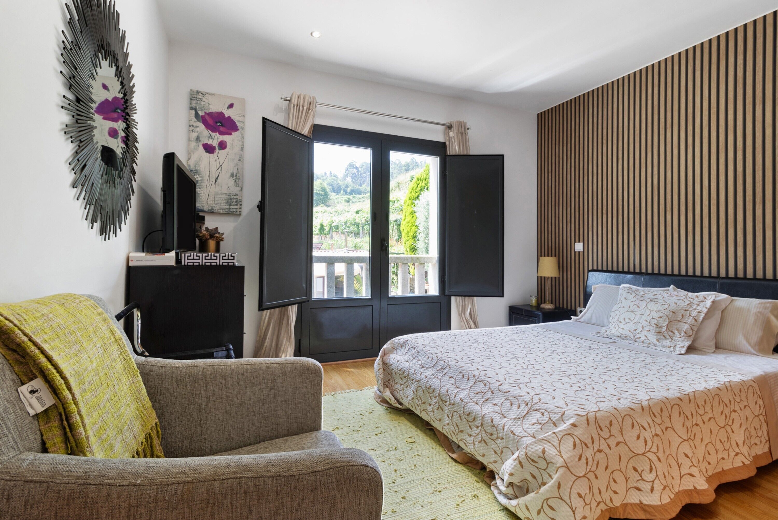 A bedroom with a bed, a chair, a TV, a window with curtains, and a wall-mounted artwork.