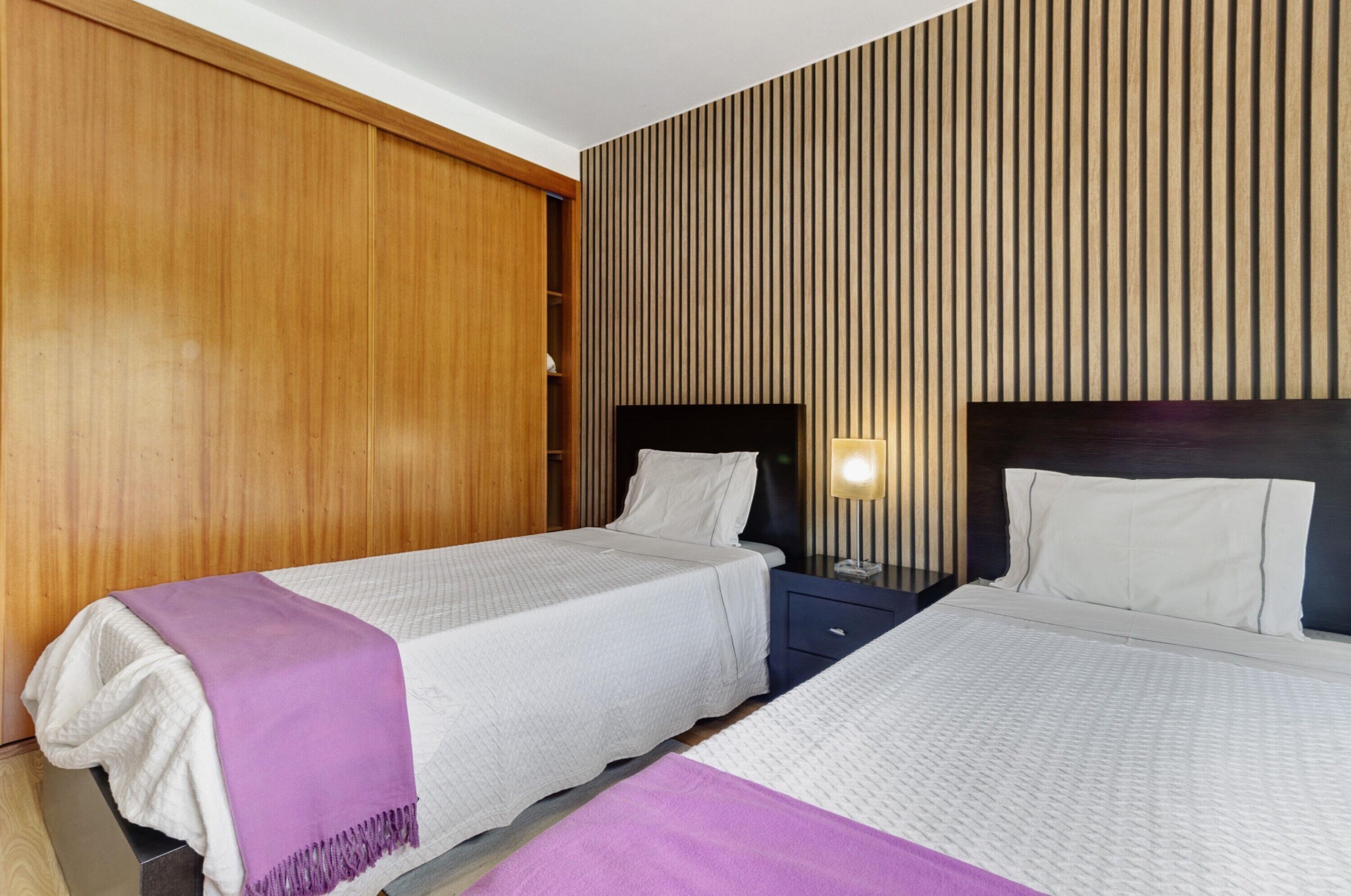 Habitación de hotel con dos camas, paneles de madera en las paredes, una mesita de noche con lámpara y un armario.