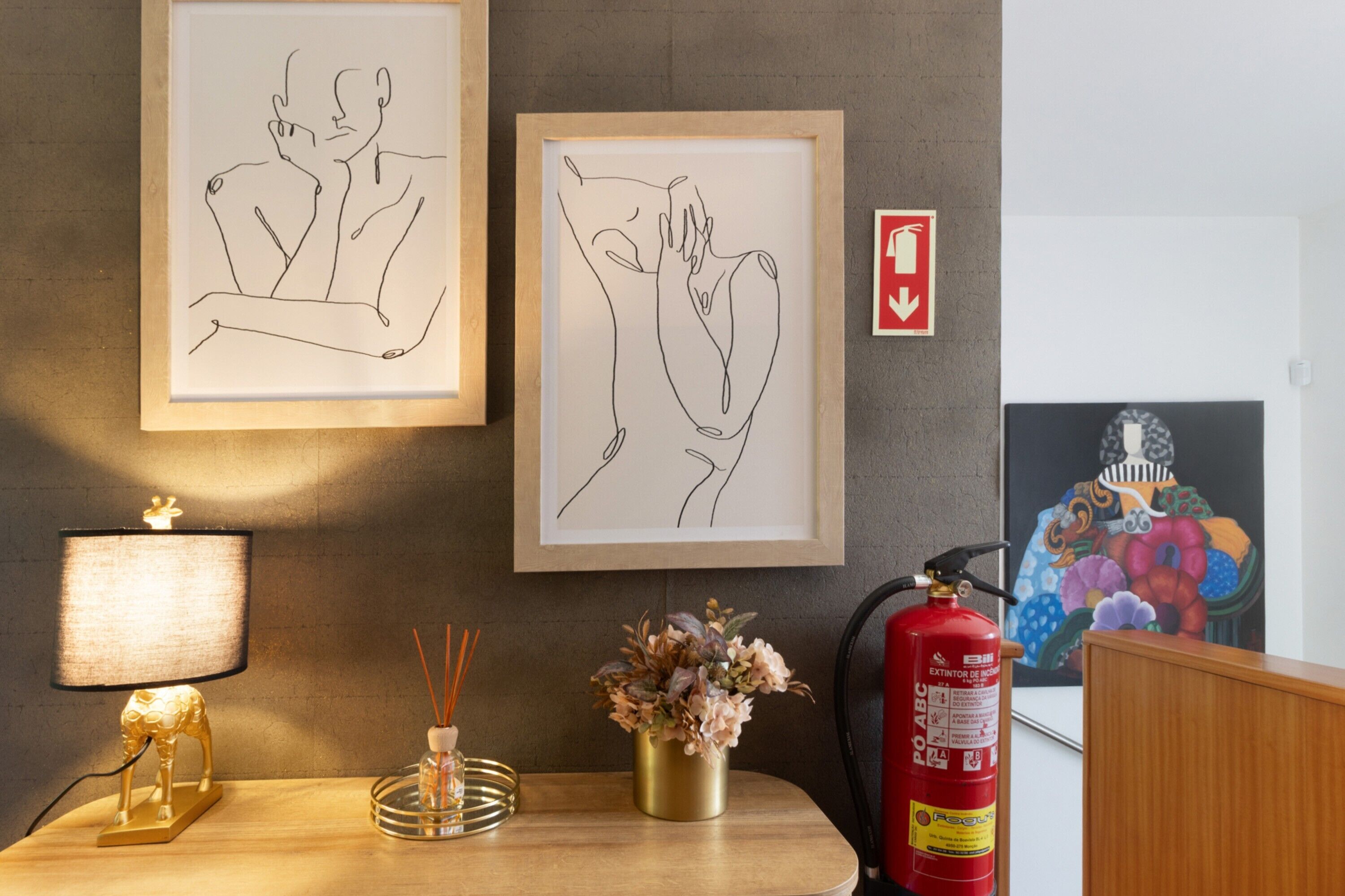 Een ingelijste tekening van twee personen, een brandblusser, een rookmelder, een decoratieve lamp en een vaas met bloemen op een houten tafel.