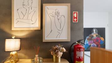 Een ingelijste tekening van twee personen, een brandblusser, een rookmelder, een decoratieve lamp en een vaas met bloemen op een houten tafel.
