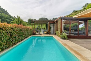 A swimming pool with a wooden deck, a covered seating area, and a view of a lush green hillside.